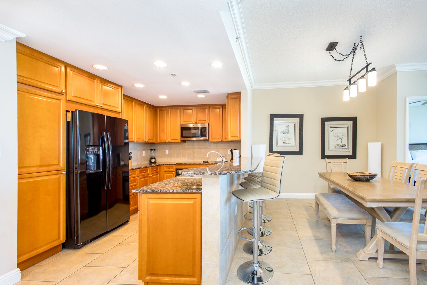 A modern kitchen with wooden cabinets, a stainless steel refrigerator, a microwave, and a granite countertop. There is a bar stool and a dining table with chairs.