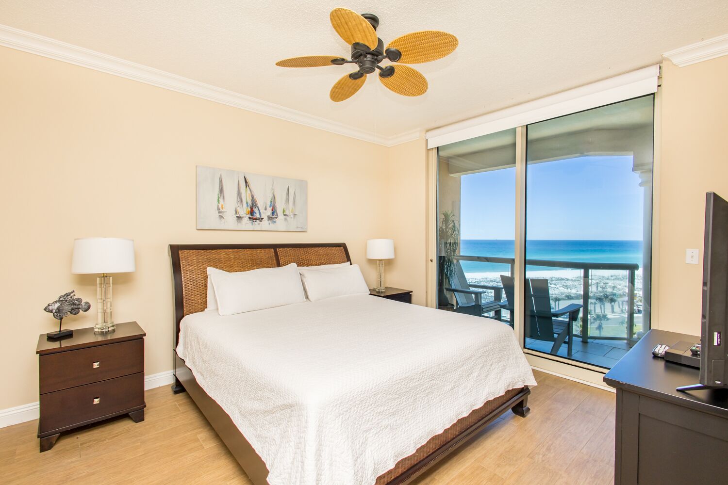 Ein Schlafzimmer mit einem Bett, Nachttischen, einer Lampe, einem Deckenventilator, einem Gemälde und einem Balkon mit Meerblick.
