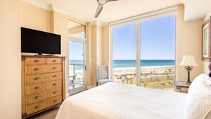 Un dormitorio con cama, televisión sobre una cómoda, ventilador de techo y una ventana grande con vista a la playa.