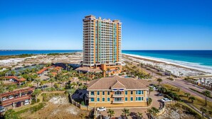 Un hôtel de grande hauteur offrant une vue sur la plage, doté de nombreux balcons et d’un parking agrémenté de palmiers.