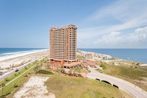 Een hoog hotel met een gebogen gevel, gelegen nabij een strand met een weg en palmbomen.