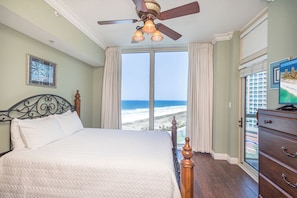 Een slaapkamer met een bed, een plafondventilator, een dressoir en een groot raam met uitzicht op het strand.