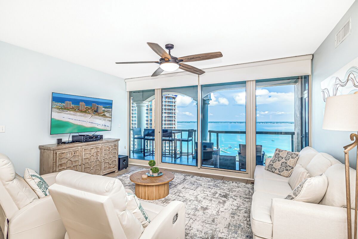 A living room with a large TV, a ceiling fan, a sofa, a coffee table, and a view of the ocean through large windows.