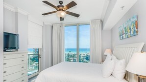 Een slaapkamer met een groot bed, een plafondventilator, een televisie en uitzicht op de stad en het strand.