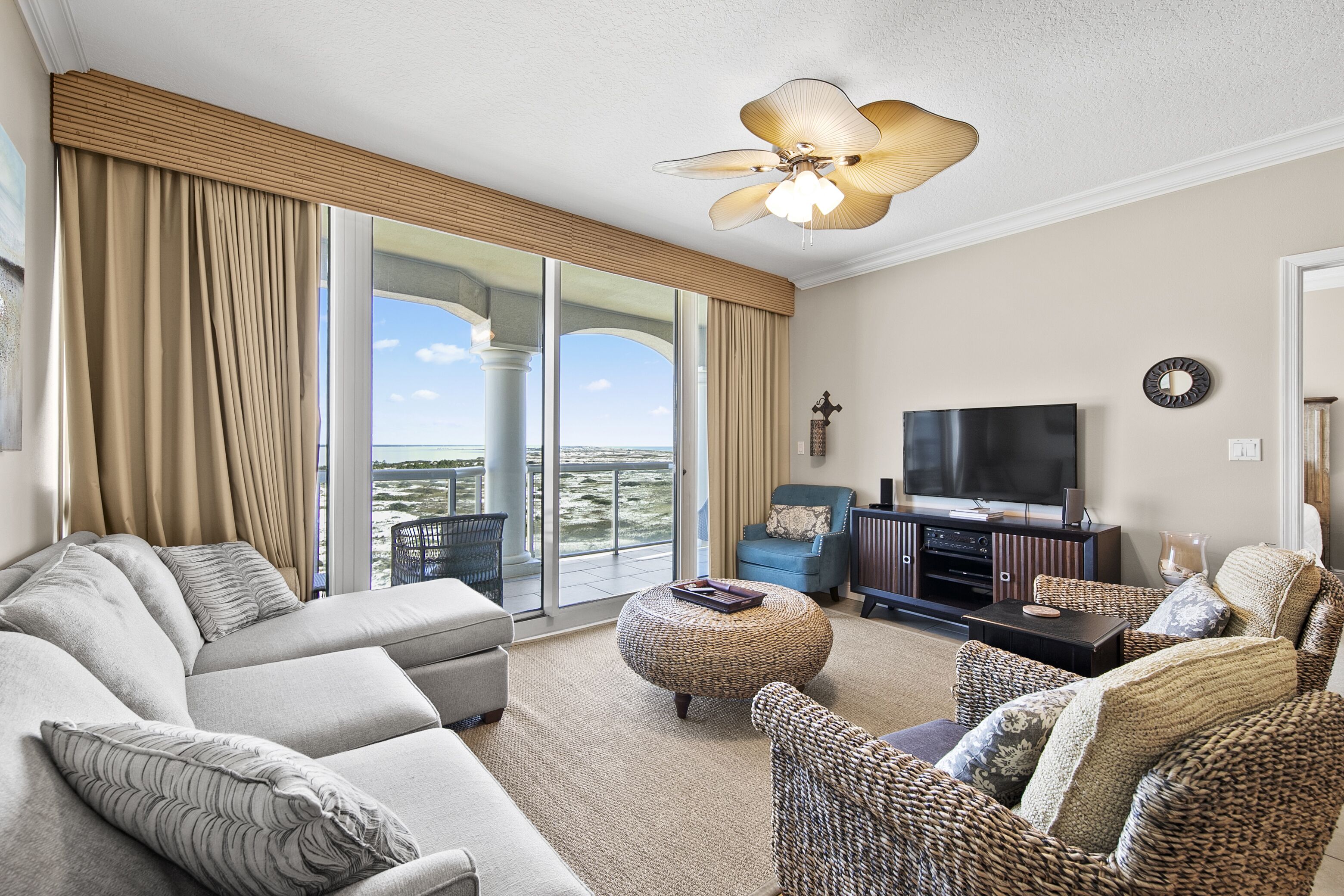 Een ruime woonkamer met een grote bank, een rieten poef, een flatscreen televisie en een balkon met uitzicht.