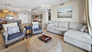 Un salon spacieux avec un canapé d’angle gris, deux fauteuils à motifs, une table basse et un coin repas comprenant une table et des chaises en bois.