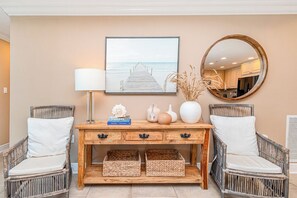 A room with a wooden console table, two wicker chairs, a framed picture of a pier, a round mirror, and wicker baskets. - P2-1506 2B Gulf View @ Portofino (Pensacola Beach)