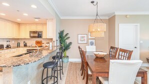 A kitchen with granite countertops, a central island with bar stools, a dining table with chairs, and a hanging light fixture. - P2-1506 2B Gulf View @ Portofino (Pensacola Beach)