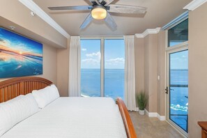 A bedroom with a large bed, a ceiling fan, a painting, and a view of the ocean through a sliding glass door.