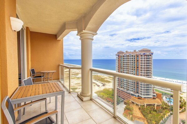 A balcony with a wooden table and chairs, overlooking a beach and a high-rise building. - P2-2006 2B Gulf View @ Portofino (Pensacola Beach)