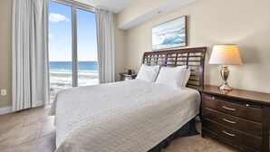 Ein Schlafzimmer mit einem großen Bett, einem Nachttisch, einer Lampe und Blick auf das Meer durch ein großes Fenster.