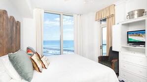 Ein Hotelzimmer mit einem großen Bett, einem Fernseher, einem Holz-Kopfteil, Blick auf den Ozean und einer Minibar.