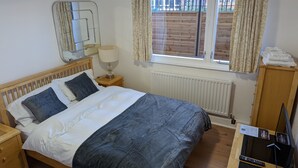 Een net opgemaakt bed met een grijze deken, een bedlampje, een houten nachtkastje en uitzicht op een bakstenen muur door een raam met gordijnen.