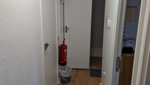 A narrow hallway with white walls, a wooden floor, a fire extinguisher, a trash bin, and a door leading to a closet. - Beautiful Independent Apartment in London, UK (London)