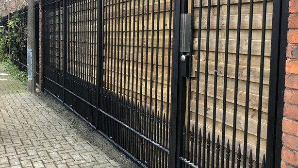 Een zwart metalen hek met verticale staven en puntige bovenkanten, naast een gebouw van baksteen met ramen.