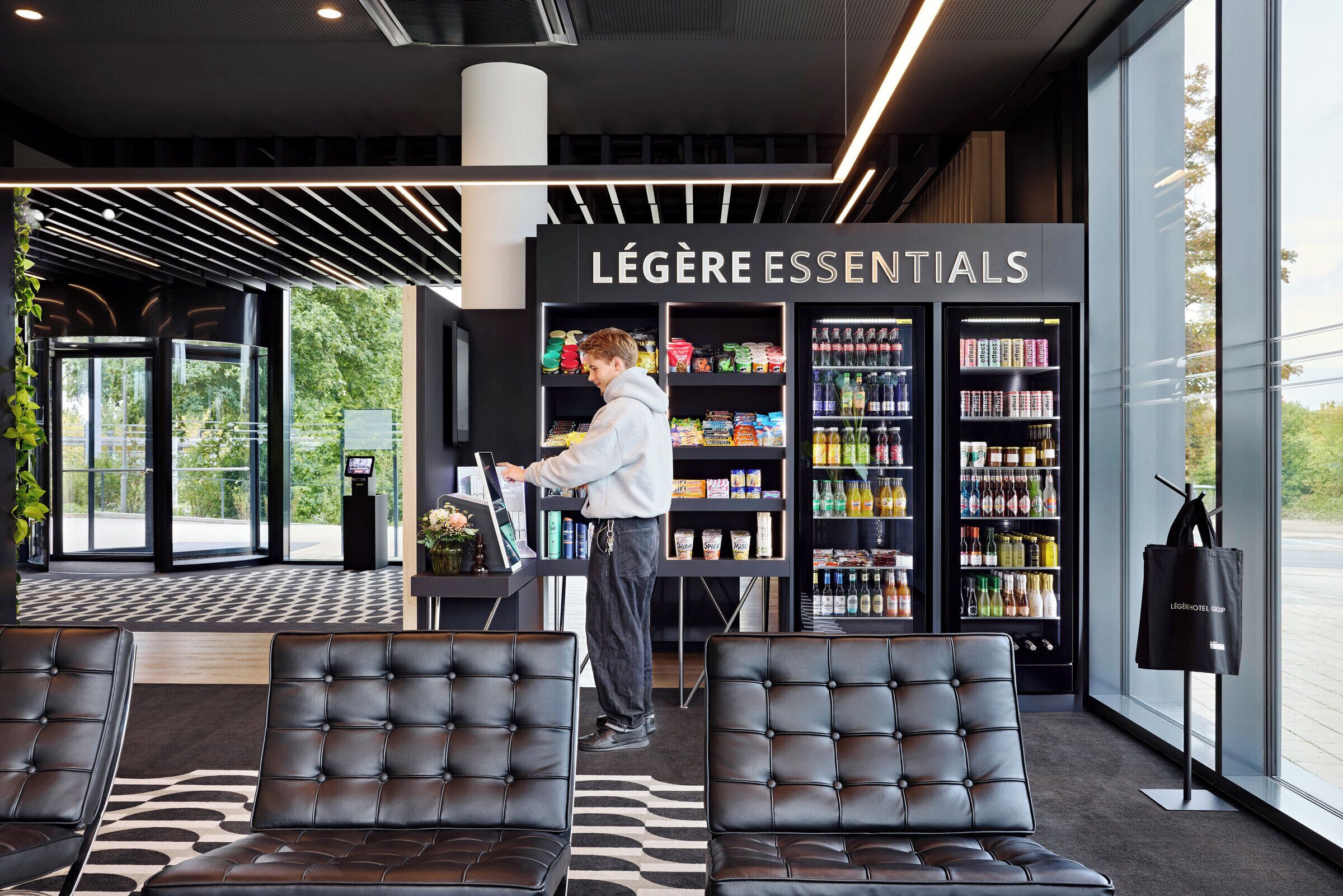 Un hall d'hôtel moderne avec des sièges en cuir, un kiosque en libre-service et un espace réfrigéré proposant une sélection de produits « Légère Essentials ».