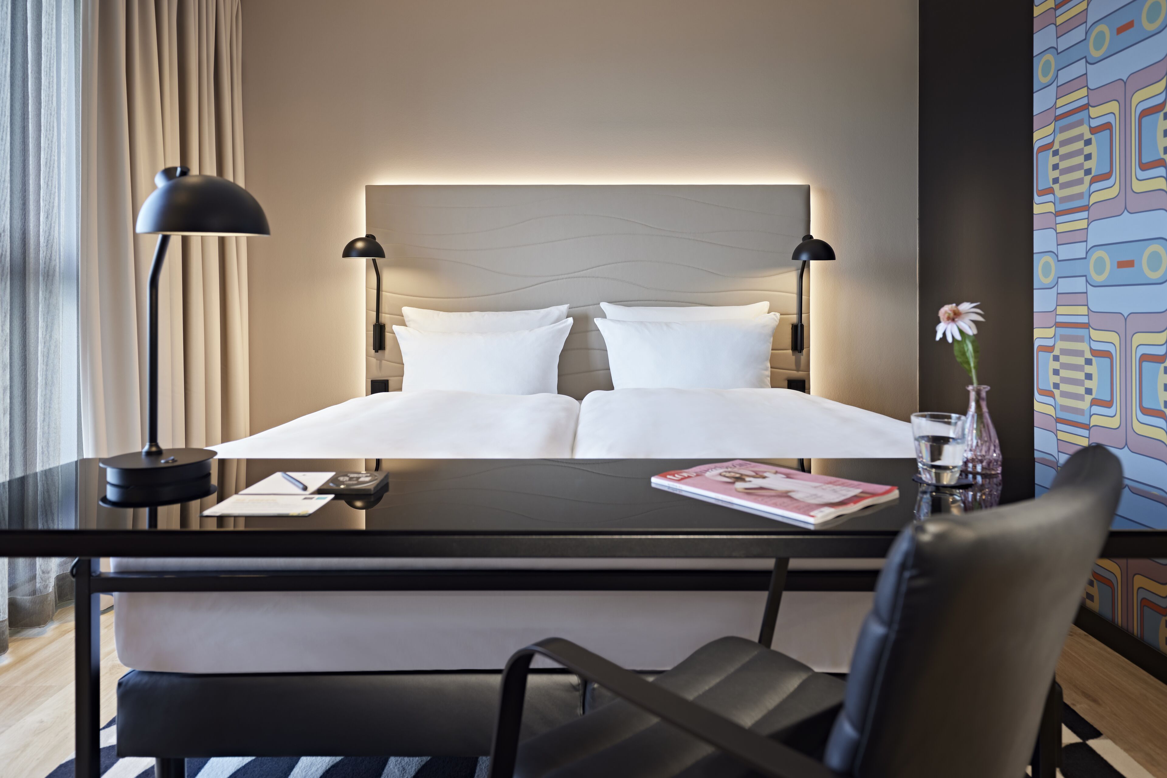 Une chambre d’hôtel avec un lit, des lampes de chevet, un bureau avec une chaise, un vase avec une fleur et un mur décoré d’un motif.
