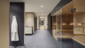 Un pasillo moderno de hotel con un muro de cristal a la derecha, una bata blanca colgada a la izquierda y una zona de sauna de madera al final.