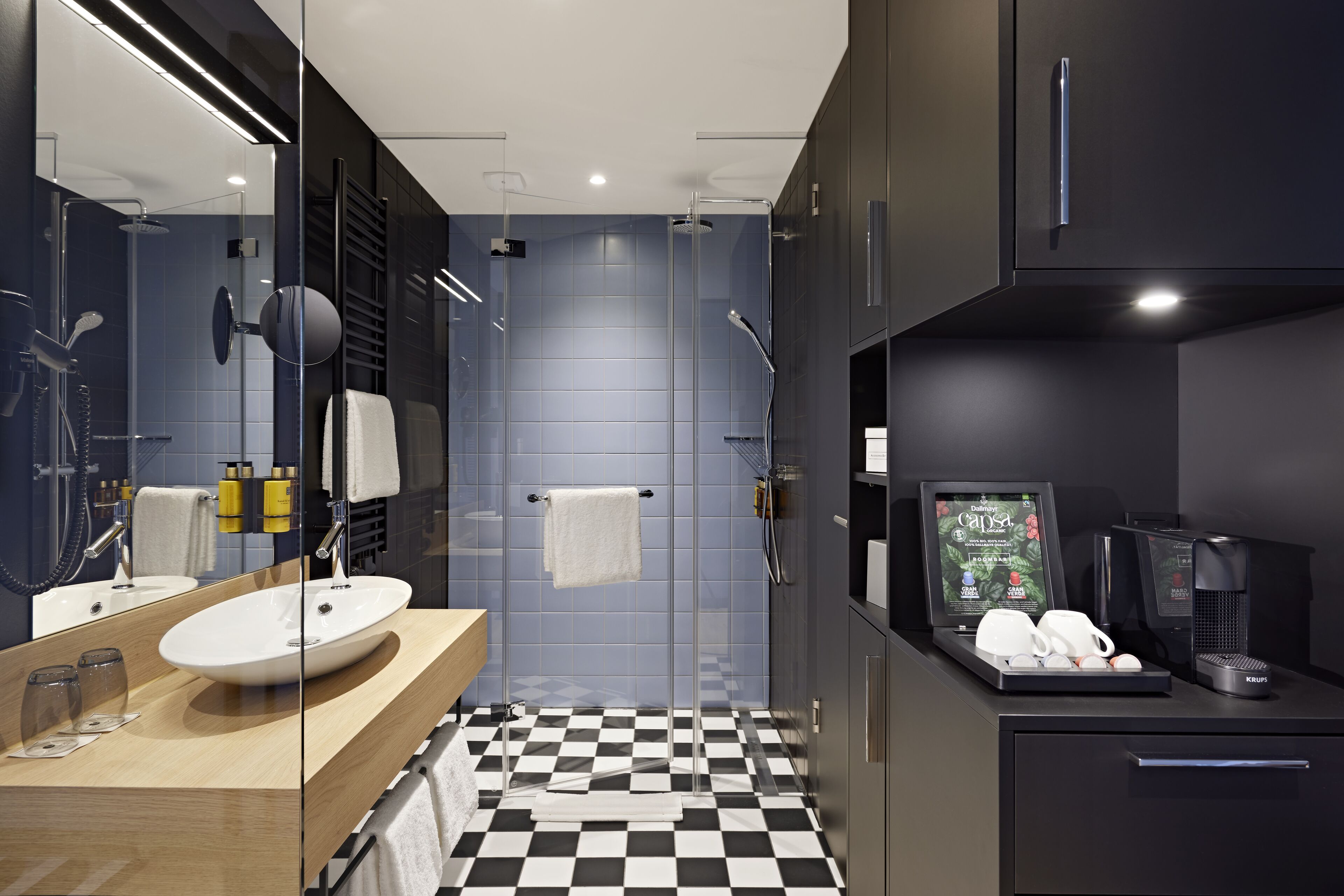 A modern bathroom with a checkered floor, a glass shower enclosure, a sink with a mirror, and a coffee machine.