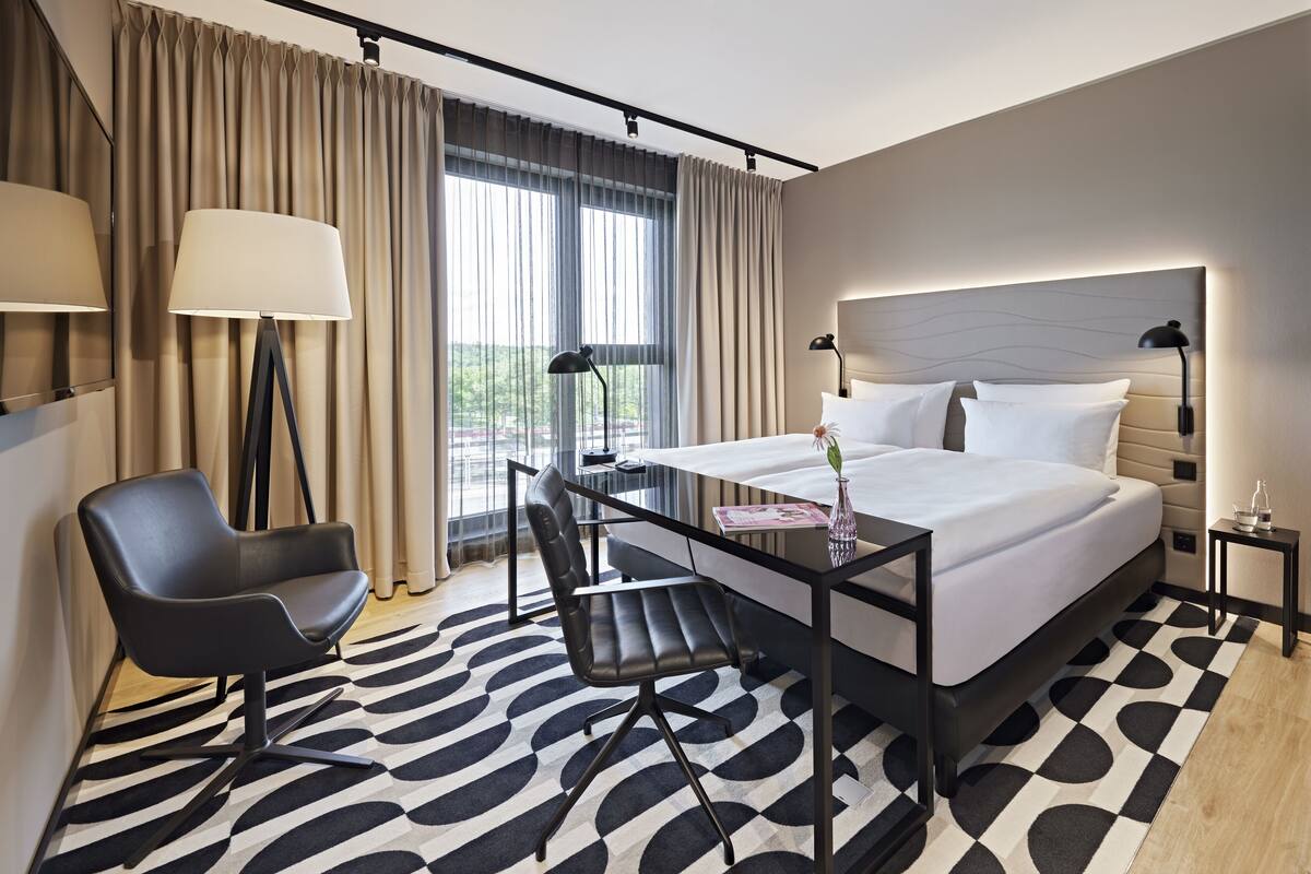 Habitación de hotel moderna con una cama grande, un escritorio con silla, una lámpara y vistas al exterior.