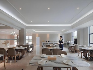 Ampla sala de jantar com várias mesas e cadeiras, uma pessoa servindo e um design de interiores moderno.