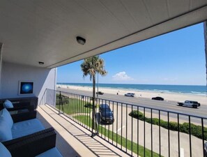 Een balkon met uitzicht op het strand en de oceaan, een palmboom en een geparkeerde auto.
