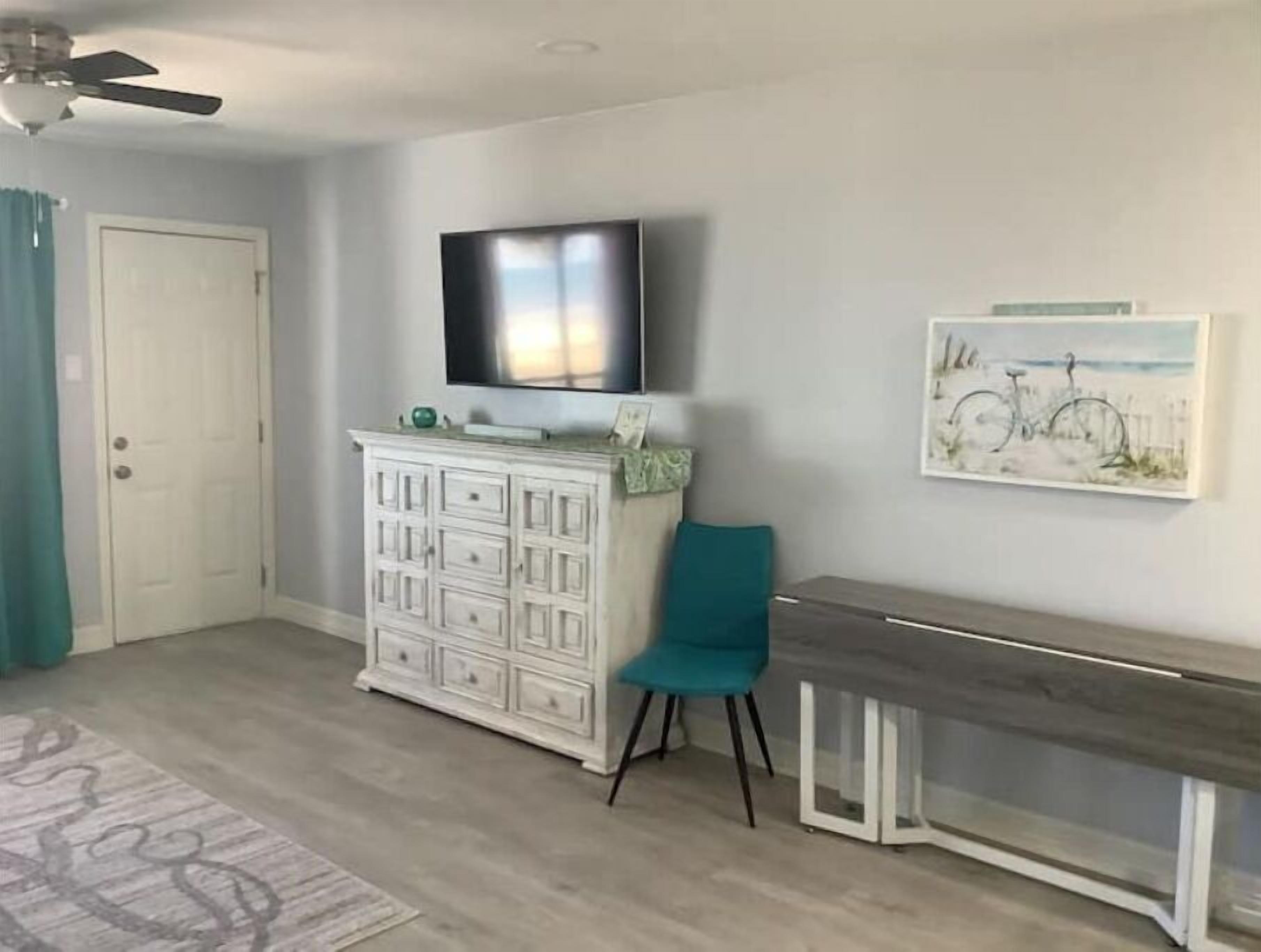 A room with a white cabinet, teal chair, and a desk with a TV mounted above.