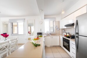 A modern kitchen with white cabinetry, a stainless steel refrigerator, a microwave, and a dining area with white chairs and a table.