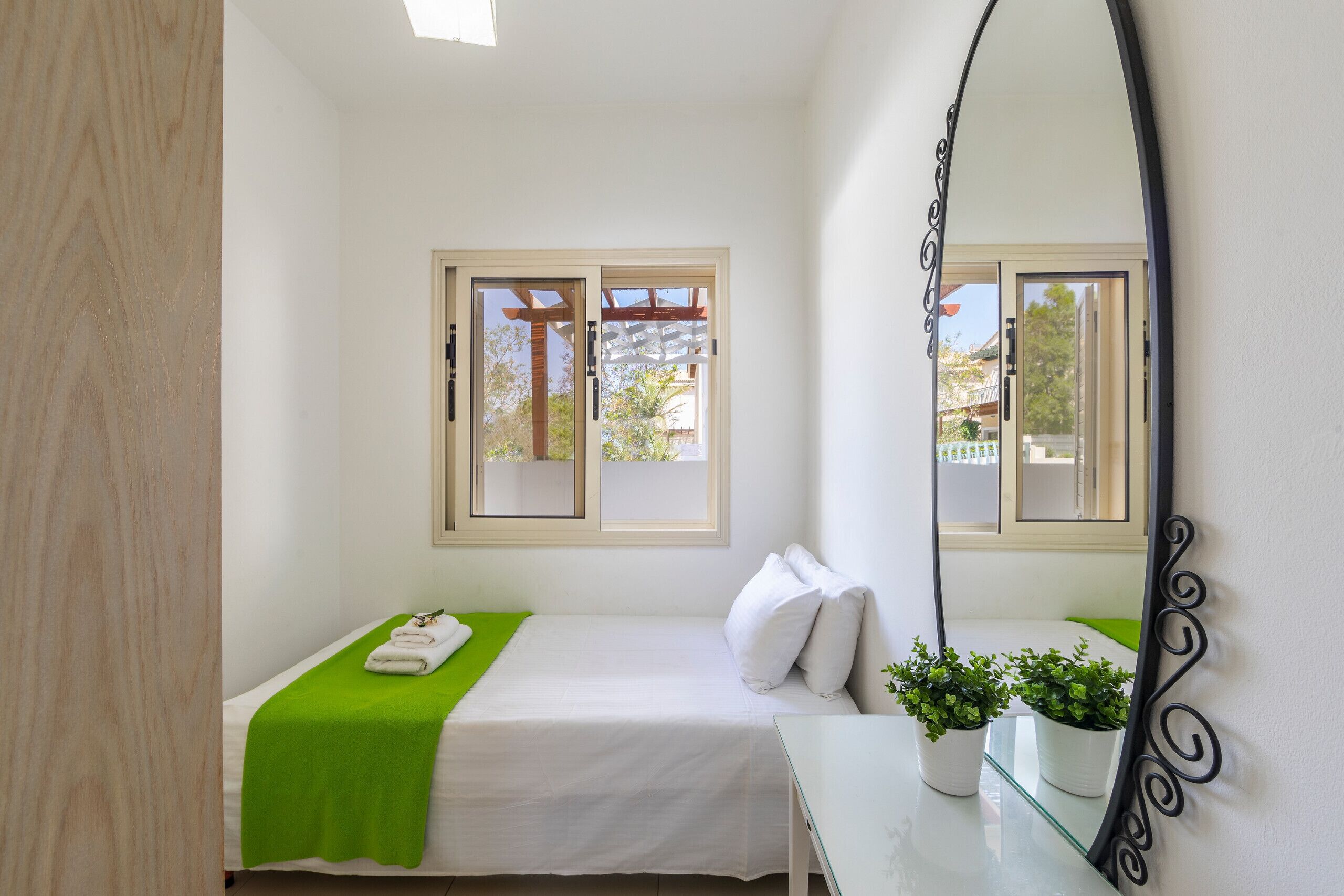 A small, clean room with a bed, a mirror, a window, and a small table with plants.