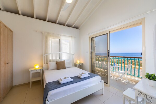 Un dormitorio con cama, mesitas de noche, un balcón con mesa y sillas, y vista al mar.