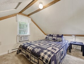 A bedroom with a sloped ceiling, a bed with a checkered bedspread, a window with blinds, an air conditioning unit, and a desk with a lamp.