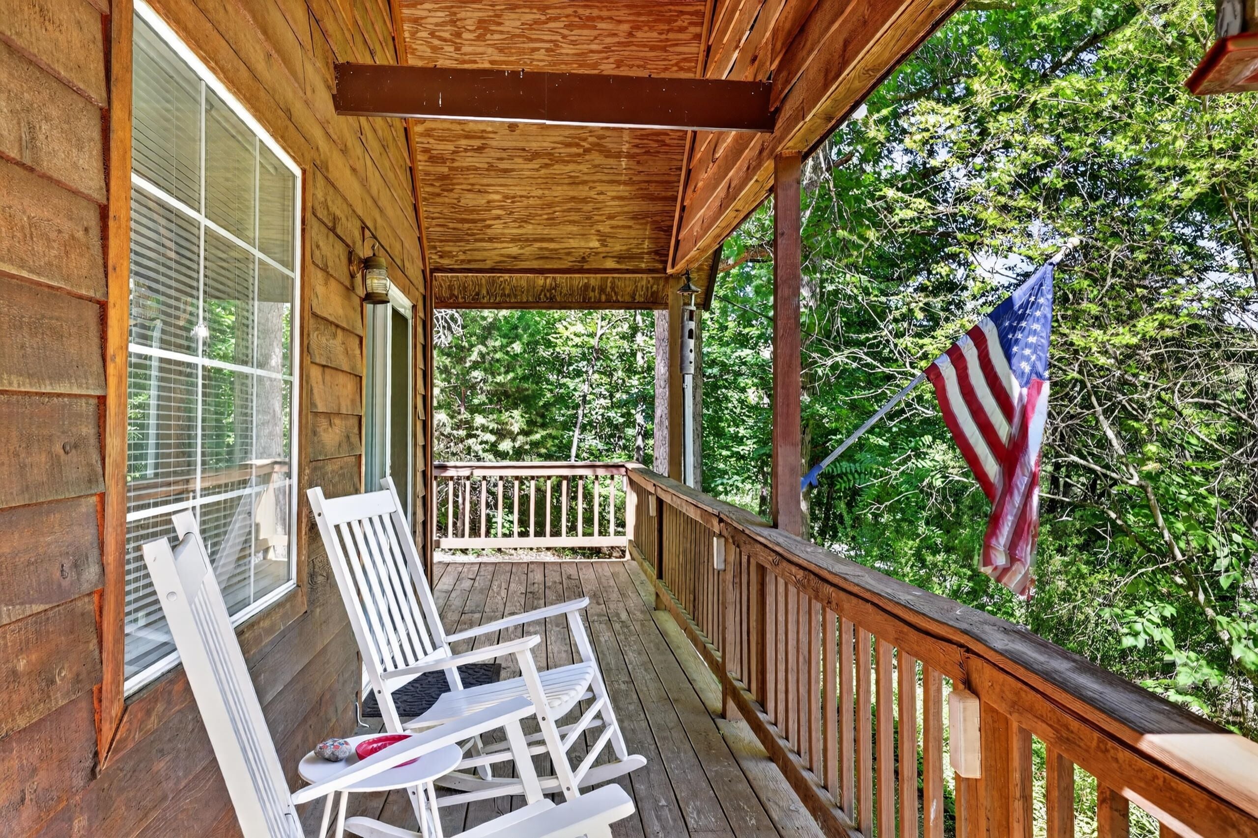A wooden porch with a railing, a flag, and two white chairs.