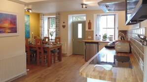 Une cuisine avec un plancher en bois, un coin repas avec une table et des chaises, un réfrigérateur et une fenêtre avec des rideaux.
