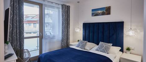 A modern bedroom with a large bed, a view of the city through a sliding door, and a cityscape painting on the wall.