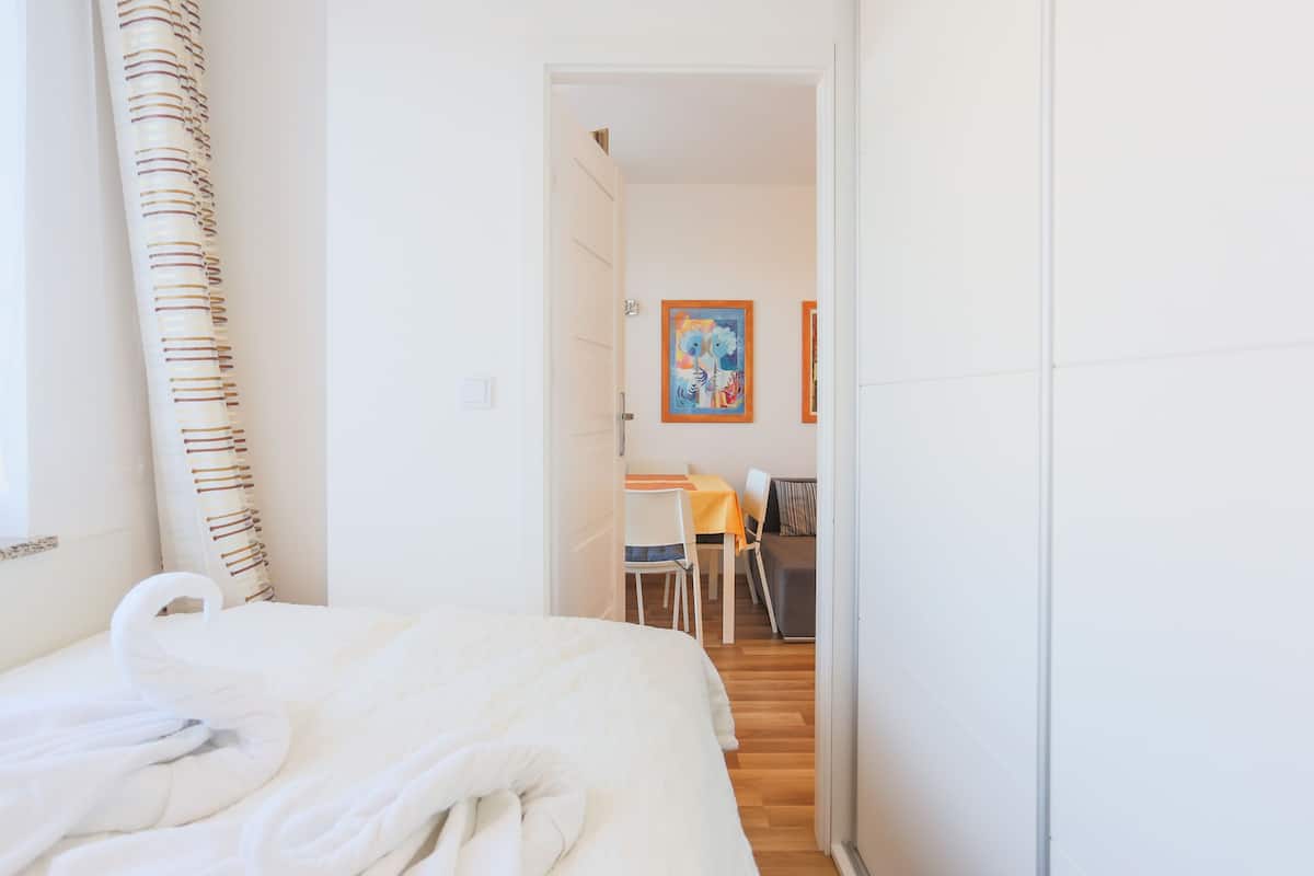 A hotel room with a bed, a shower curtain, a painting on the wall, and a dining area with a table and chairs.