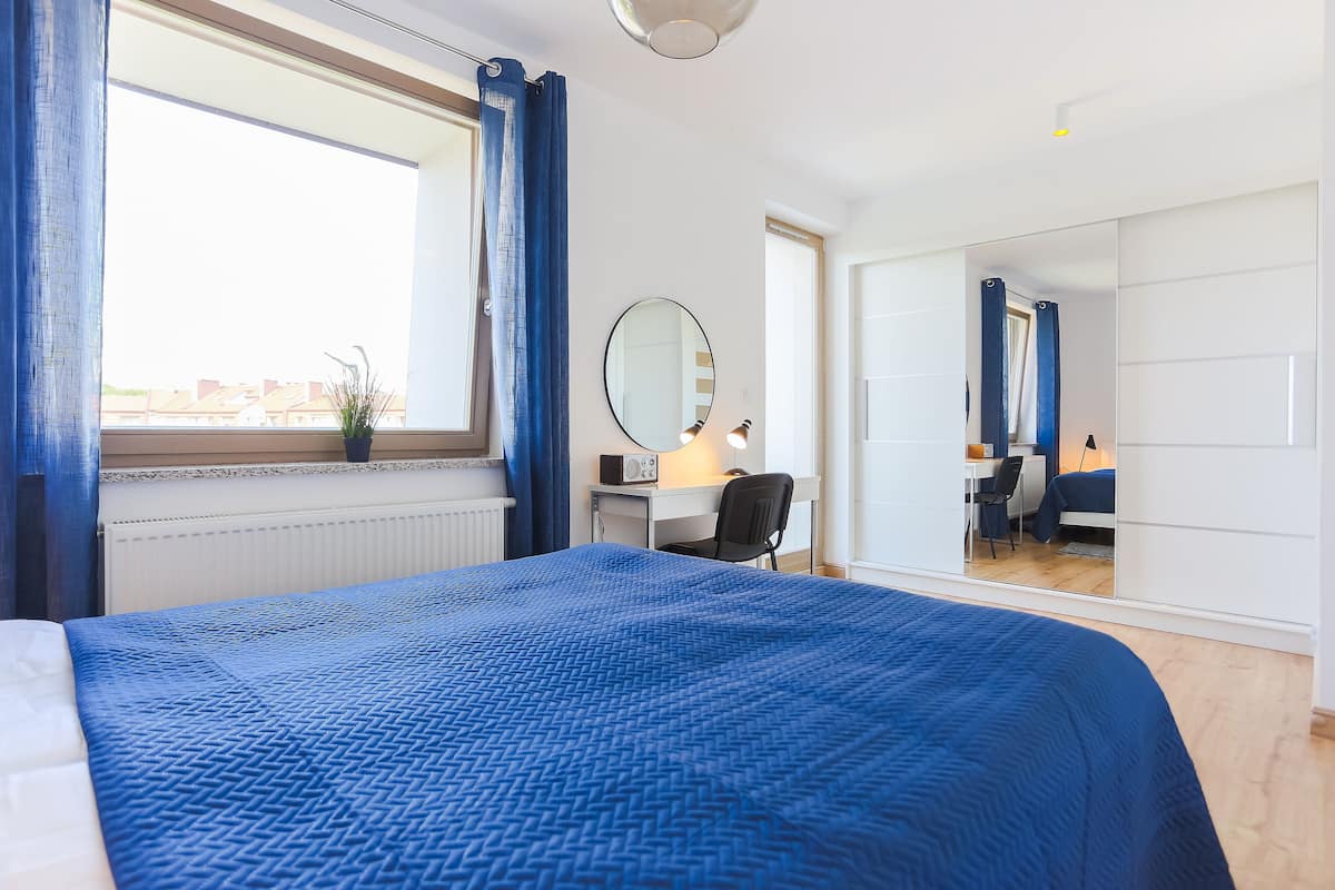 A modern hotel room with a large bed, a desk, a chair, a mirror, and a view of the city through the window.