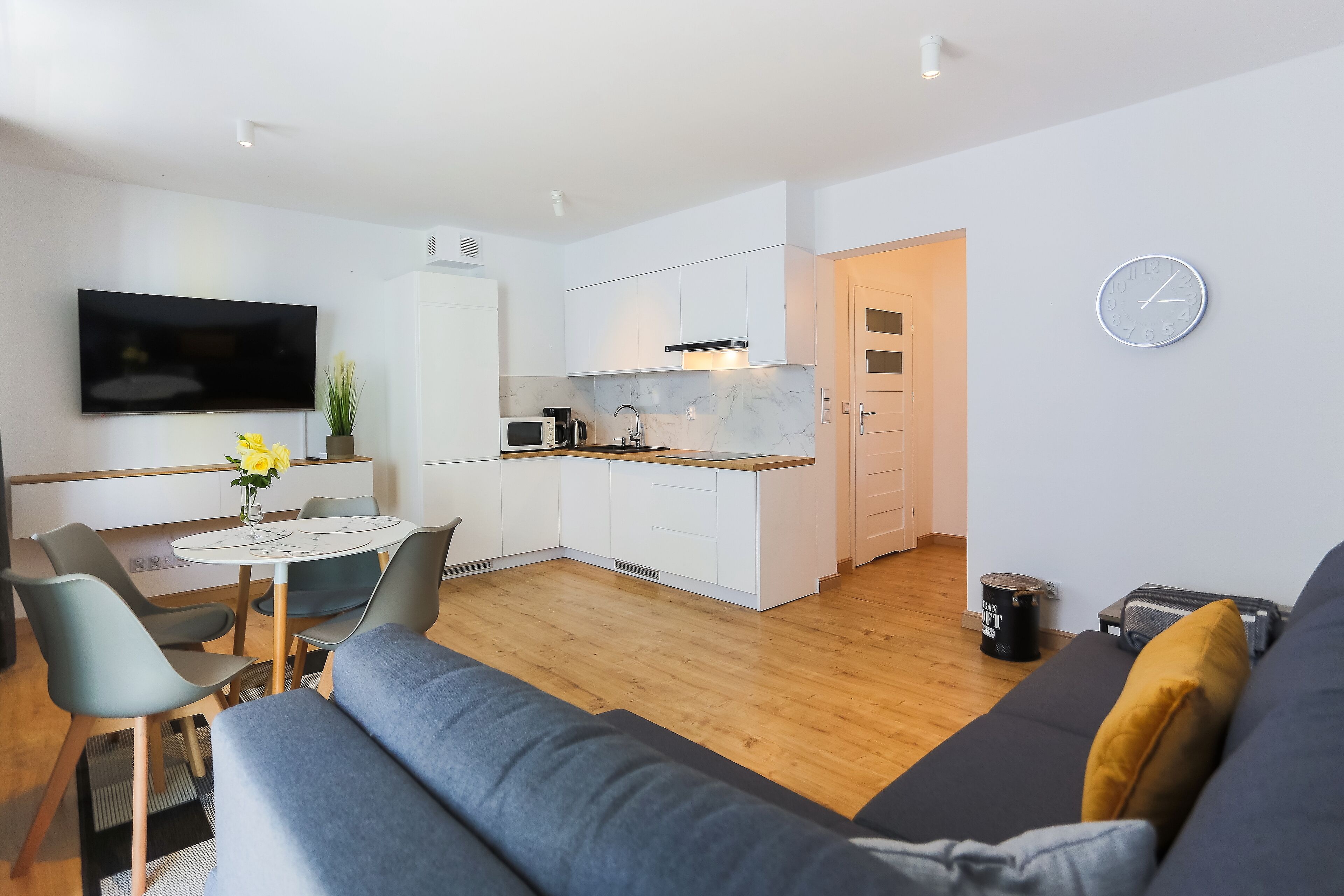 A modern living room with a sofa, a dining table with chairs, a TV, and a kitchen area with a microwave and stove.