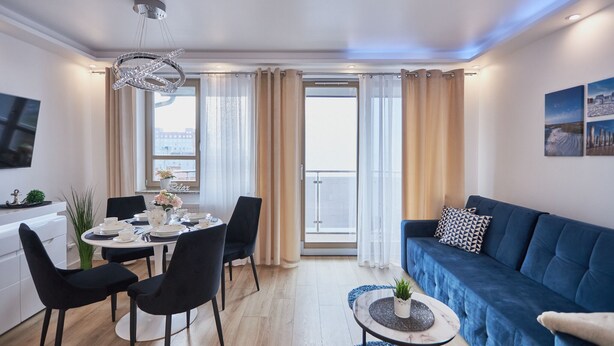 A modern living room with a blue sofa, a dining table set for four, and a large window with curtains. - Apartamenty Swinoujscie K Rozy Wiatrow 2 (Swinoujscie)