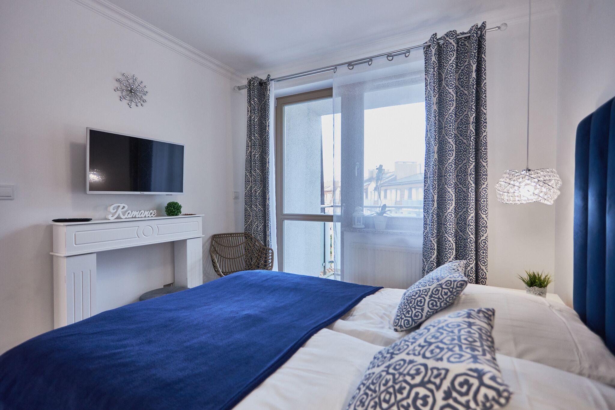 Un dormitorio con cama, televisión sobre un tocador blanco, ventana con cortinas estampadas y vista a la ciudad.