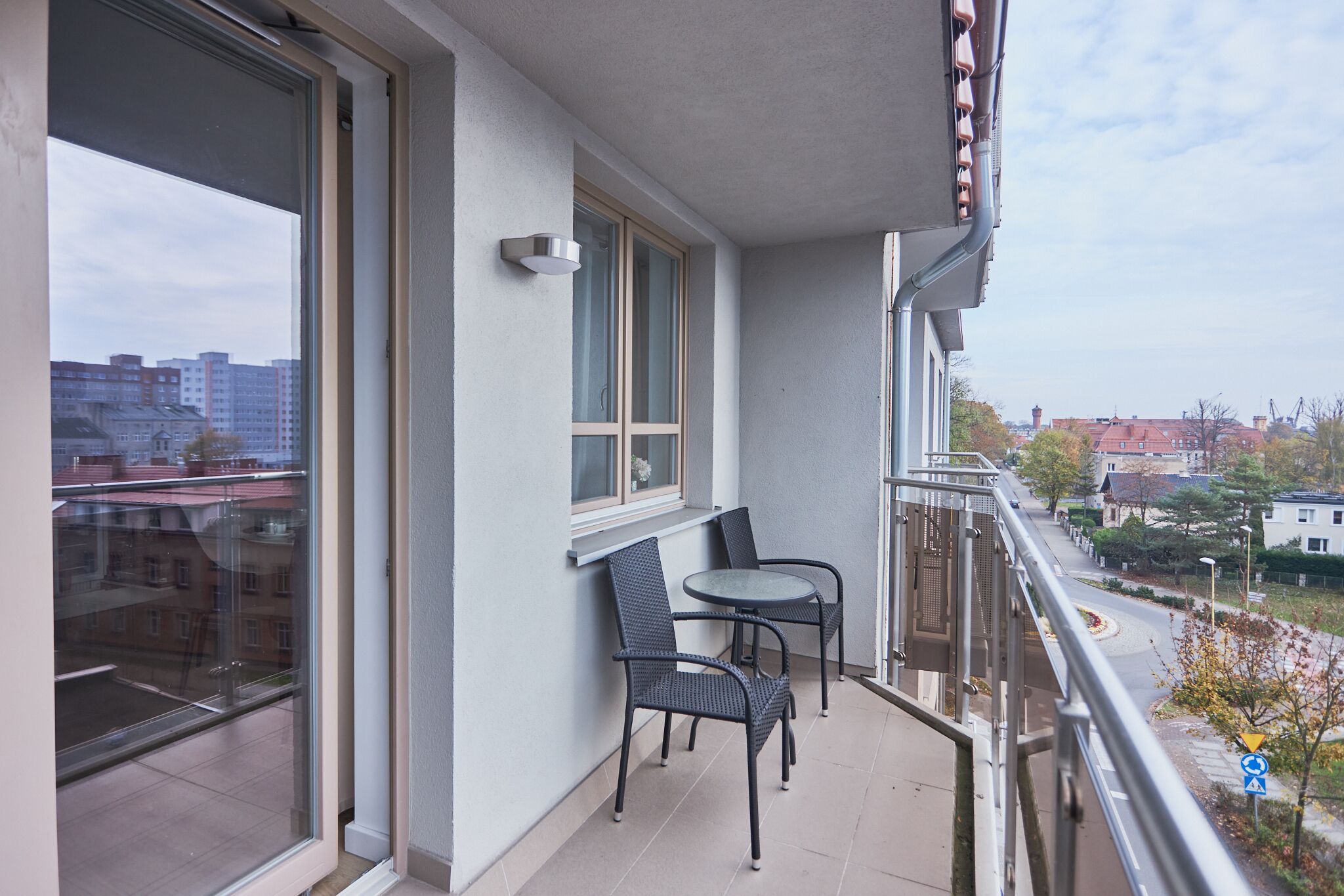 A balcony with two chairs and a table, large windows, and a view of buildings and trees.