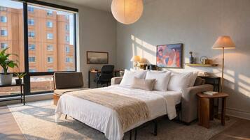 A modern hotel room with a large bed, a desk, a chair, a potted plant, and a view of the city.
