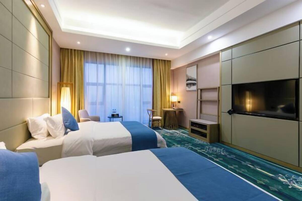 A hotel room with two beds, a desk, a chair, a TV, and a large window with curtains.