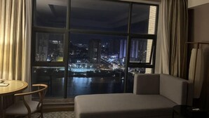 A hotel room with a large window offering a nighttime city view, a sofa, a small table, and a chair.