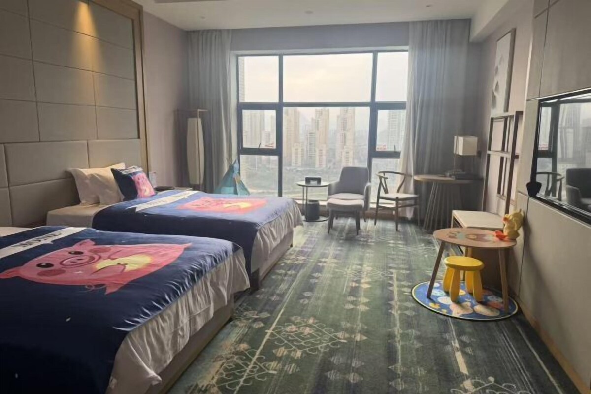 A hotel room with two beds, a desk, a chair, a small table, and a view of the cityscape.