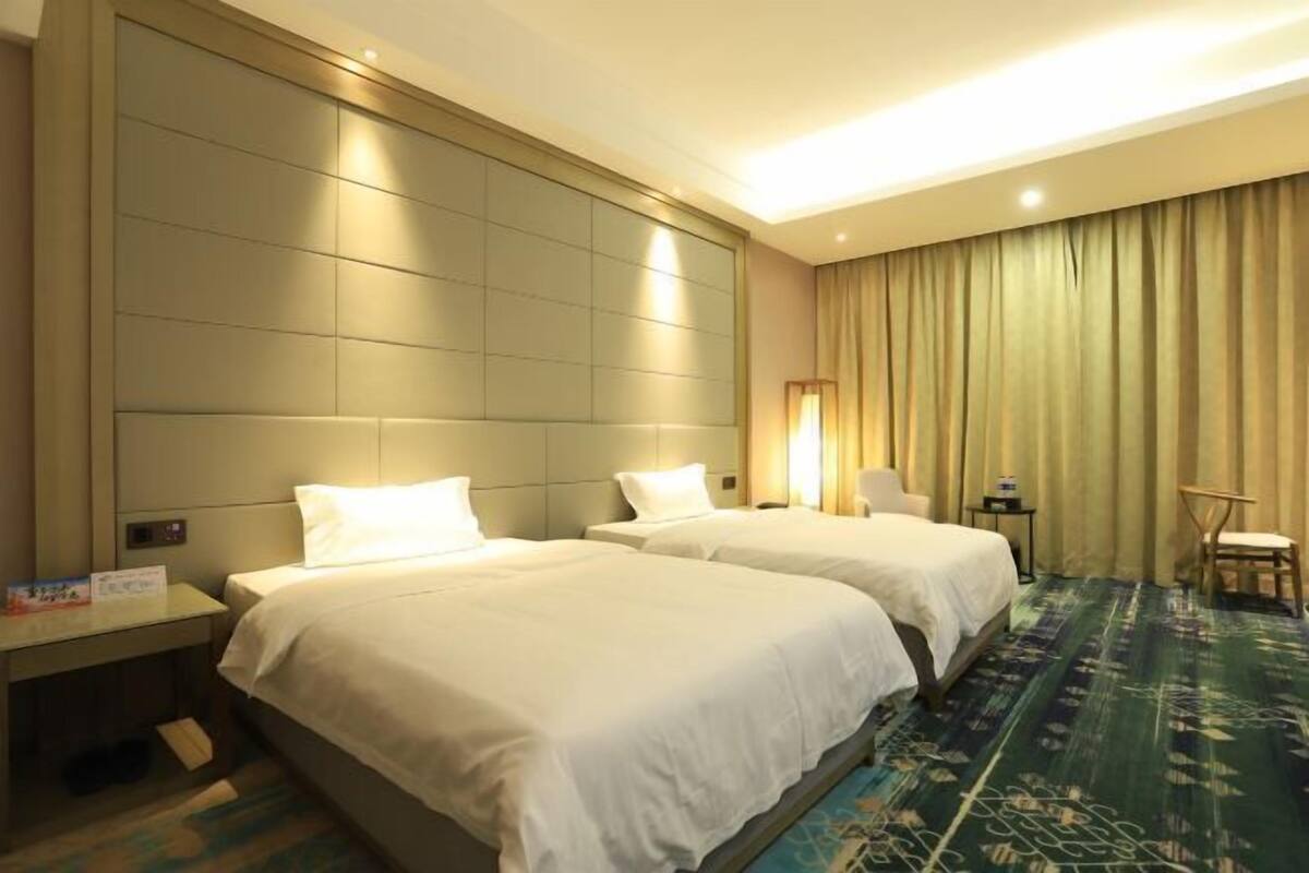 A hotel room with two beds, a large window with curtains, a small table with a lamp, and a carpeted floor.