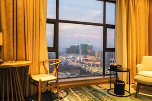 Habitación de hotel con un ventanal grande que ofrece vistas a la ciudad, una mesa redonda, dos sillas, una mesita auxiliar y un cubo de basura.