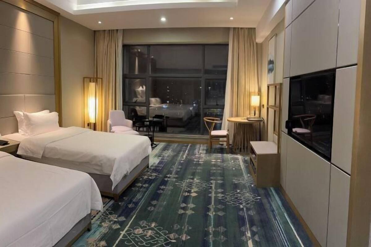 A hotel room with two beds, a desk, a chair, a TV, and a large window.