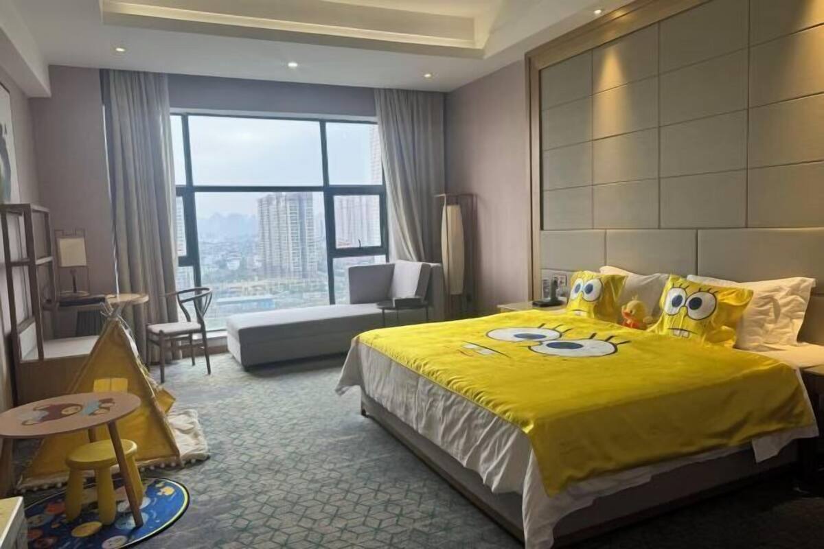 A hotel room with a large bed, a desk, a chair, a small table, and a window with a city view.
