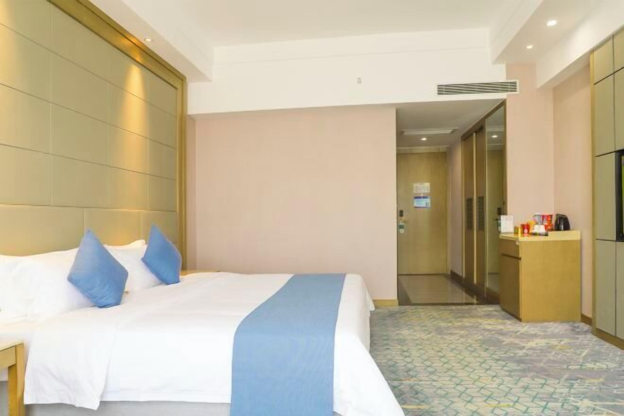 A hotel room with a bed, a bedside table, a desk, a minibar, and a bathroom visible in the background.
