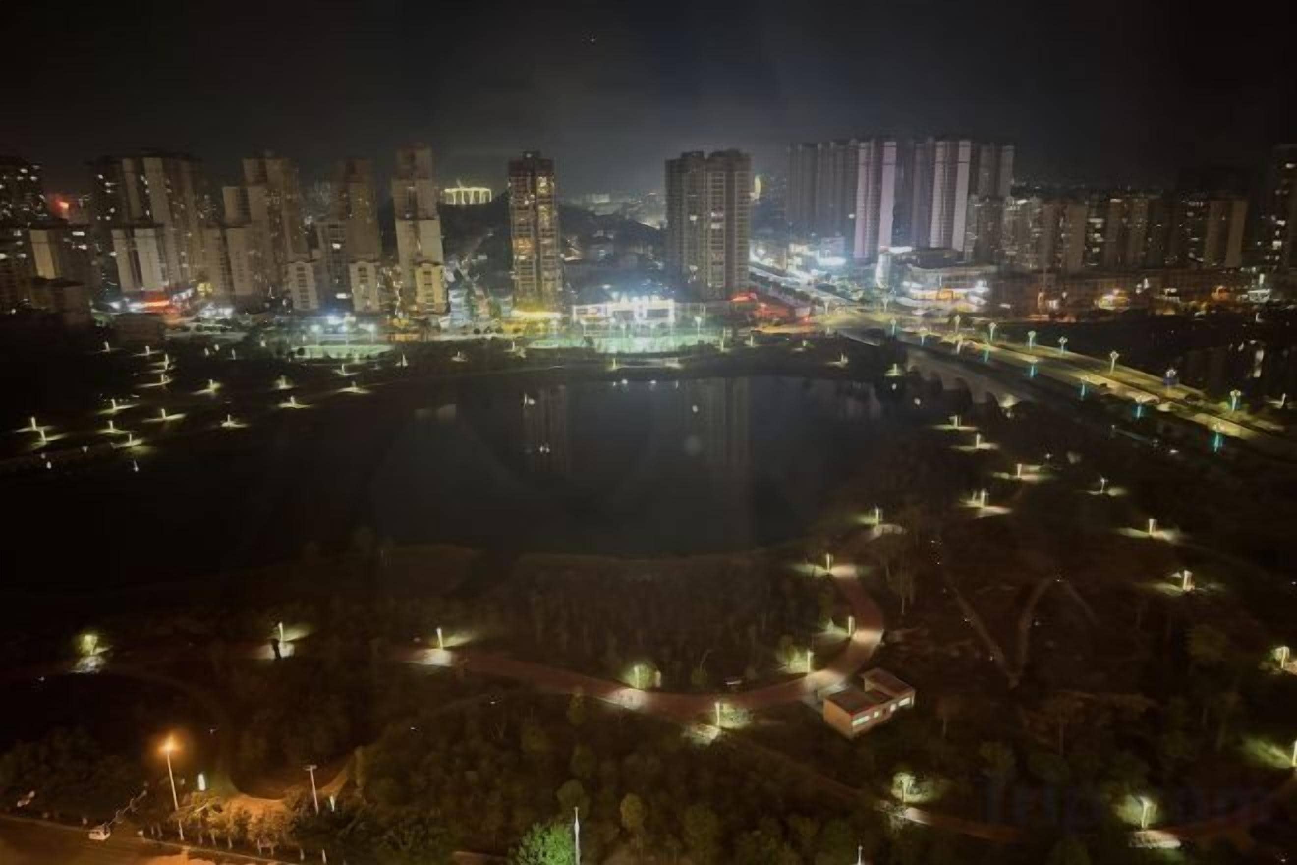 A nighttime cityscape with a body of water reflecting lights, surrounded by buildings and a park area with pathways.